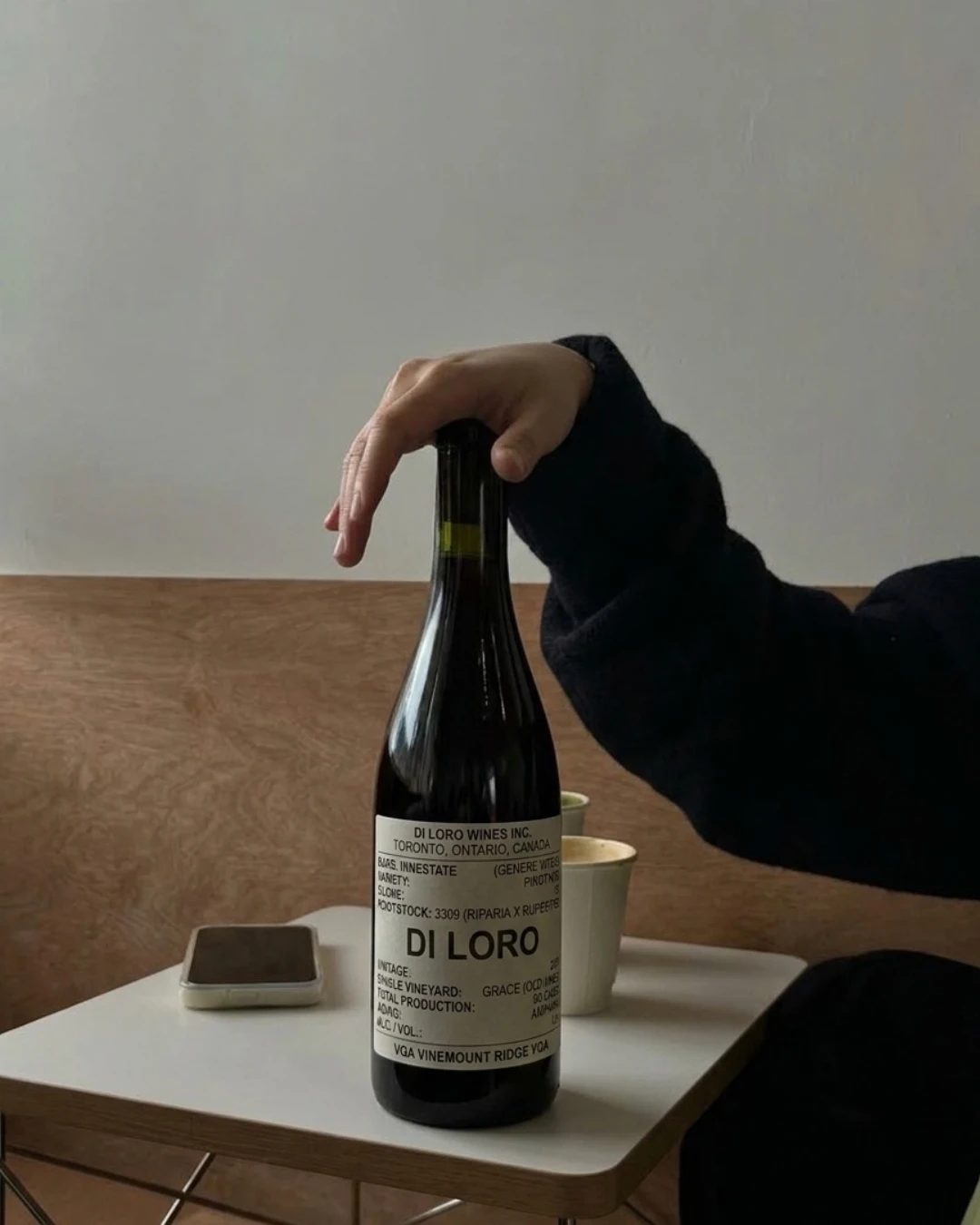 Une main posée sur une bouteille de Di Loro, un rouge VQA de Vinemount Ridge en Ontario, sur une petite table blanche