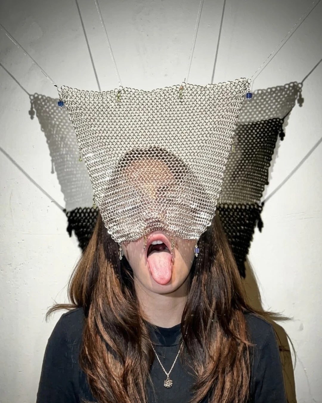 A woman with a chainmail veil over her face and her tongue out, against a white wall