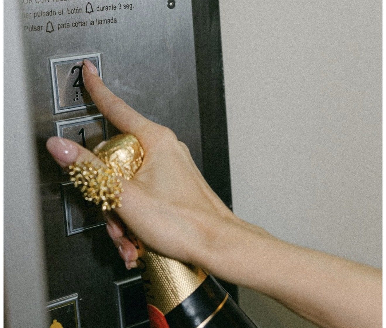 Une main tenant une bouteille de mousseux appuie sur le bouton du deuxième étage dans un ascenseur
